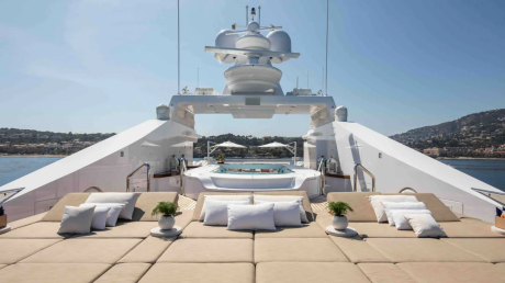 A luxurious yacht with plush seating in the foreground is anchored in a calm bay with a lush, green hillside in the background.