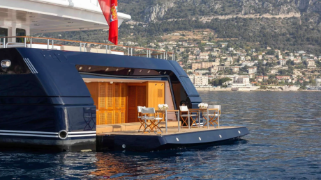 A luxurious yacht with an open deck featuring a dining set is anchored in calm blue water with a hillside town and mountains in the background.