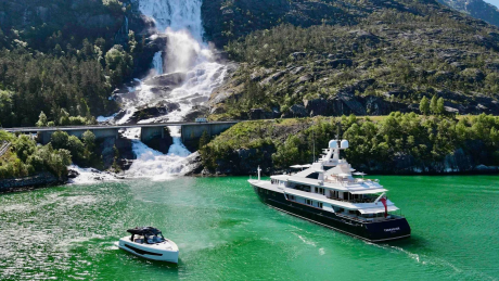 A large yacht and a smaller boat sail on vibrant green water in front of a dramatic waterfall cascading down a rocky mountainside.
