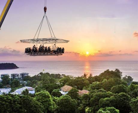 A suspended dining platform with people enjoying a meal hovers over a lush green landscape and a tranquil ocean at sunset.