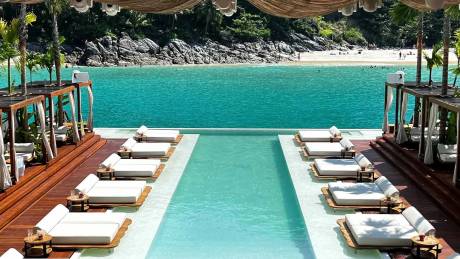 Rows of white lounge chairs surround a bright blue infinity pool in the foreground, with a sandy beach and turquoise ocean stretching into the background.