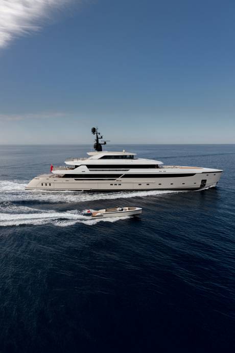 A large white yacht cruises through dark blue water, trailed by a smaller boat, under a clear blue sky.
