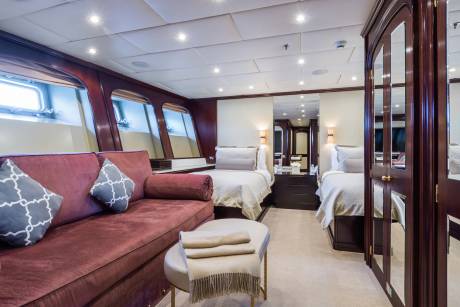 A plush maroon sofa with patterned pillows sits in the foreground of a well-lit cabin with two beds and mirrored closets in the background.