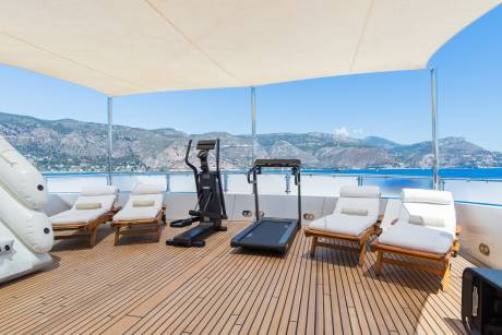 A sun-drenched deck with lounge chairs and exercise equipment faces a picturesque coastline under a clear blue sky.