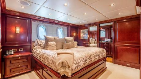 A luxurious yacht bedroom features a large bed with patterned bedding in the foreground, while the background reveals mirrored closets and a glimpse into an ensuite bathroom.