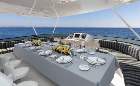 A long dining table set for a meal on a yacht's deck is in the foreground, with the vast blue ocean and clear sky in the background.
