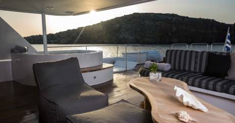 A luxurious yacht deck with a hot tub and seating area in the foreground overlooks a tranquil bay at sunset.