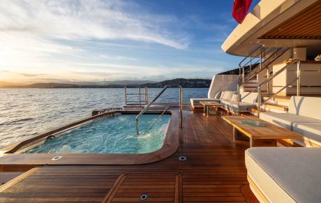 A bubbling jacuzzi and plush seating area on a yacht's deck overlook a calm sea and distant coastline under a soft sunset sky.