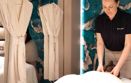 Two plush white robes hang in the foreground, while a masseuse works on a client in the background against a vibrant, textured wall.