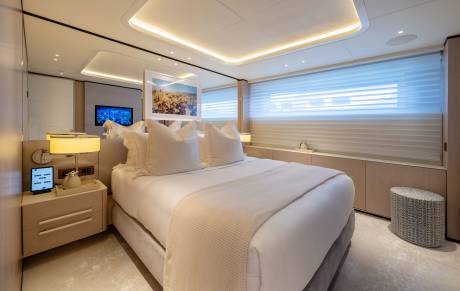 A luxurious yacht bedroom features a plush king-sized bed in the foreground and a large window with blinds and a television in the background.