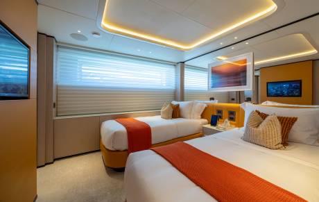 Two beds with orange throws are in the foreground, while a large window with blinds and a mounted television are in the background.