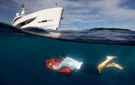 A man in a suit swims underwater on a red scooter, with a large white yacht sailing above him in the blue ocean.
