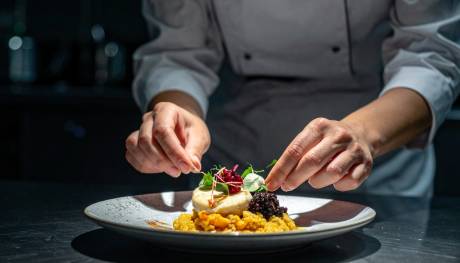 A chef's hands artfully garnish a beautifully plated dish of food in the foreground, with a blurred kitchen background behind them.