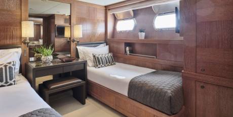 Two neatly made twin beds with patterned pillows and quilted blankets fill the foreground, while the background features wood-paneled walls, porthole windows, and a television.
