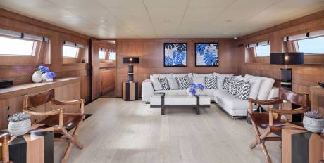 Two wooden chairs with striped side tables are in the foreground, while a large white sectional sofa with blue and white patterned pillows sits in the background, facing two framed blue floral artworks on a wood-paneled wall.