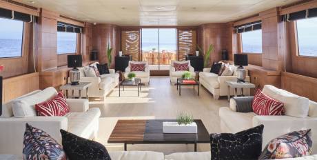 Plush white sofas with decorative pillows fill the foreground of a luxurious yacht lounge, while a bright beach scene is visible through large windows in the background.