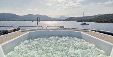 A bubbling hot tub on a yacht's deck overlooks a calm bay with distant, hazy mountains and a sailboat.