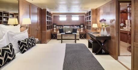 A luxurious bedroom with a large bed in the foreground and a desk and seating area in the background, all surrounded by wooden paneling and bookshelves.
