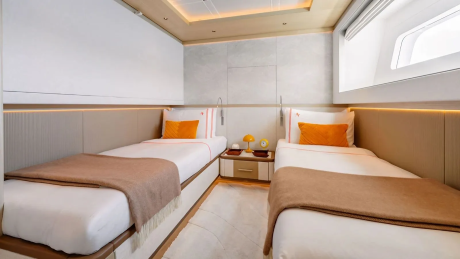 Two neatly made twin beds with orange pillows and brown throws occupy the foreground, while a nightstand with a lamp and a window are visible in the background.