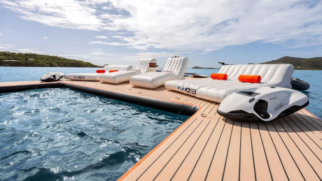 Inflatable loungers and a jet ski rest on a floating dock in the foreground, with a calm blue ocean and lush green islands in the background.