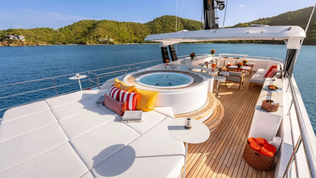 A luxurious yacht with a hot tub and lounge area in the foreground is anchored in a calm bay with lush green hills in the background.
