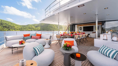 Plush white seating arrangements and a dining table are set on a yacht's deck, with a lush, green, hilly coastline visible in the background under a bright blue sky.