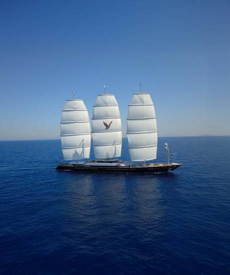 A majestic three-masted sailing yacht with full white sails glides across a calm, deep blue ocean under a clear, bright sky.