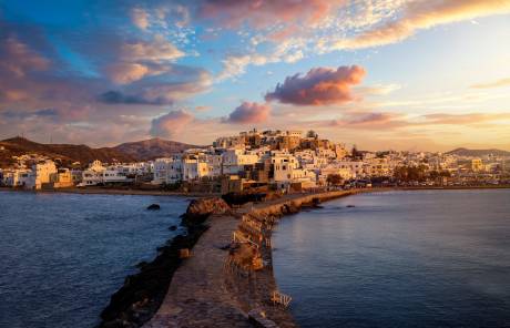 A stone pathway leads to a whitewashed village nestled on a hill overlooking the sea, under a colorful sunset sky.