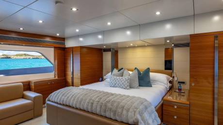 A luxurious yacht bedroom with a large bed and plush pillows in the foreground, and a window overlooking a turquoise ocean and distant islands in the background.