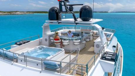 A luxurious yacht with a hot tub in the foreground cruises through clear turquoise waters with a distant, low-lying island in the background.
