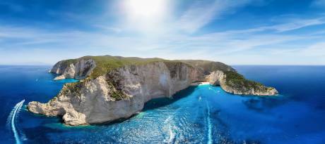 Speedboats create white wakes in the crystal-clear blue water surrounding a lush, green island with dramatic white cliffs under a bright, sunny sky.