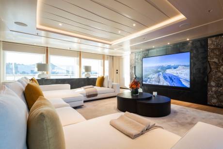A spacious, modern living area with a large sectional sofa in the foreground and a large television displaying a scenic landscape in the background.