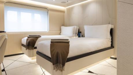 Two neatly made twin beds with brown throws are in the foreground, while a large window with blinds and a textured wall are in the background.