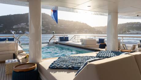 Plush lounge seating and a sparkling pool occupy the foreground, while a sun-drenched coastline with buildings and hills forms the background.