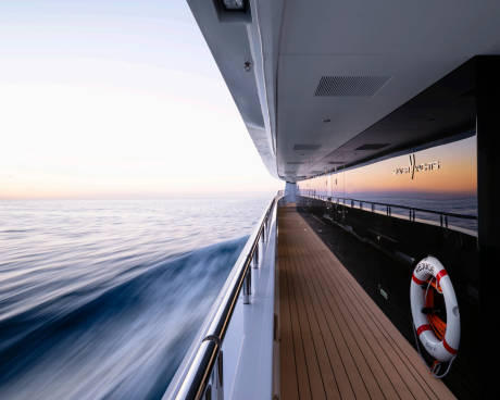A wooden deck with a life preserver runs along the side of a yacht as it cruises through calm ocean waters under a soft, hazy sky.