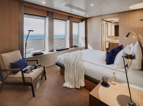 A luxurious yacht cabin with a comfortable armchair in the foreground and a large bed facing windows with a view of the ocean in the background.