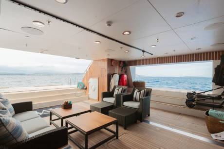 A luxurious yacht deck with comfortable seating and coffee tables in the foreground overlooks a calm blue ocean and distant islands in the background.