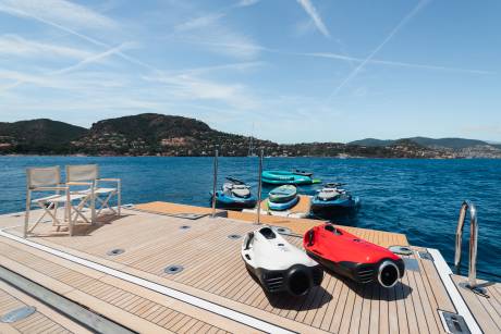 Two underwater scooters rest on a wooden deck in the foreground, with jet skis and an inflatable raft docked behind them against a backdrop of blue water and a distant, tree-covered coastline under a clear sky.
