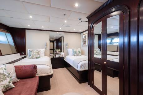 A luxurious yacht cabin features two twin beds with floral pillows in the foreground, and a large mirrored wardrobe and a window with a view of the water in the background.