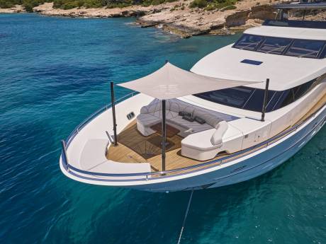 A luxurious white yacht with a shaded seating area in the foreground floats on clear turquoise water near a rocky, tree-lined coastline in the background.