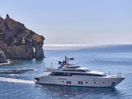 A sleek white yacht cruises through calm blue waters in the foreground, with a dramatic rocky cliff face and the shimmering sea stretching to the horizon in the background.