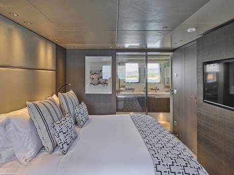 A modern bedroom with a plush bed in the foreground and a glass-enclosed bathroom with a view of the sea in the background.