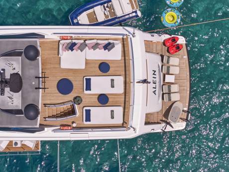 A luxurious yacht with comfortable seating and sun loungers is anchored in clear turquoise water, with a small dinghy and inflatable tubes nearby.