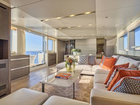 A spacious, modern yacht living area with a large sectional sofa and coffee table in the foreground, and expansive windows overlooking the ocean in the background.