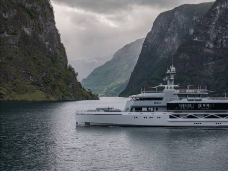 A sleek, modern yacht sails on calm water between towering, verdant cliffs under a cloudy sky.