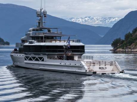 A large white yacht named "BOLD" cruises through calm water with snow-capped mountains in the background.