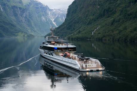 A large white yacht with a helipad sails through a calm, reflective fjord, flanked by steep, green mountains.