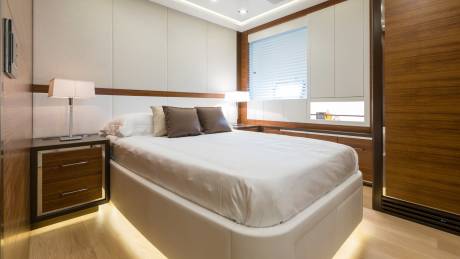 A luxurious bedroom with a white bed and bedside table in the foreground, and a window with blinds and wooden cabinets in the background.