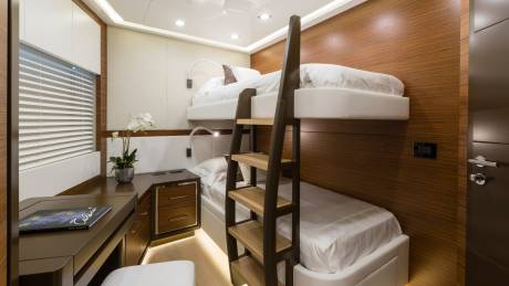 A bunk bed with white linens is in the foreground, while a wooden-paneled wall and a desk with blinds are in the background.