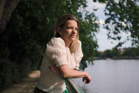 A woman in a white blouse leans on a railing by a path, looking out at a calm river with trees in the background.